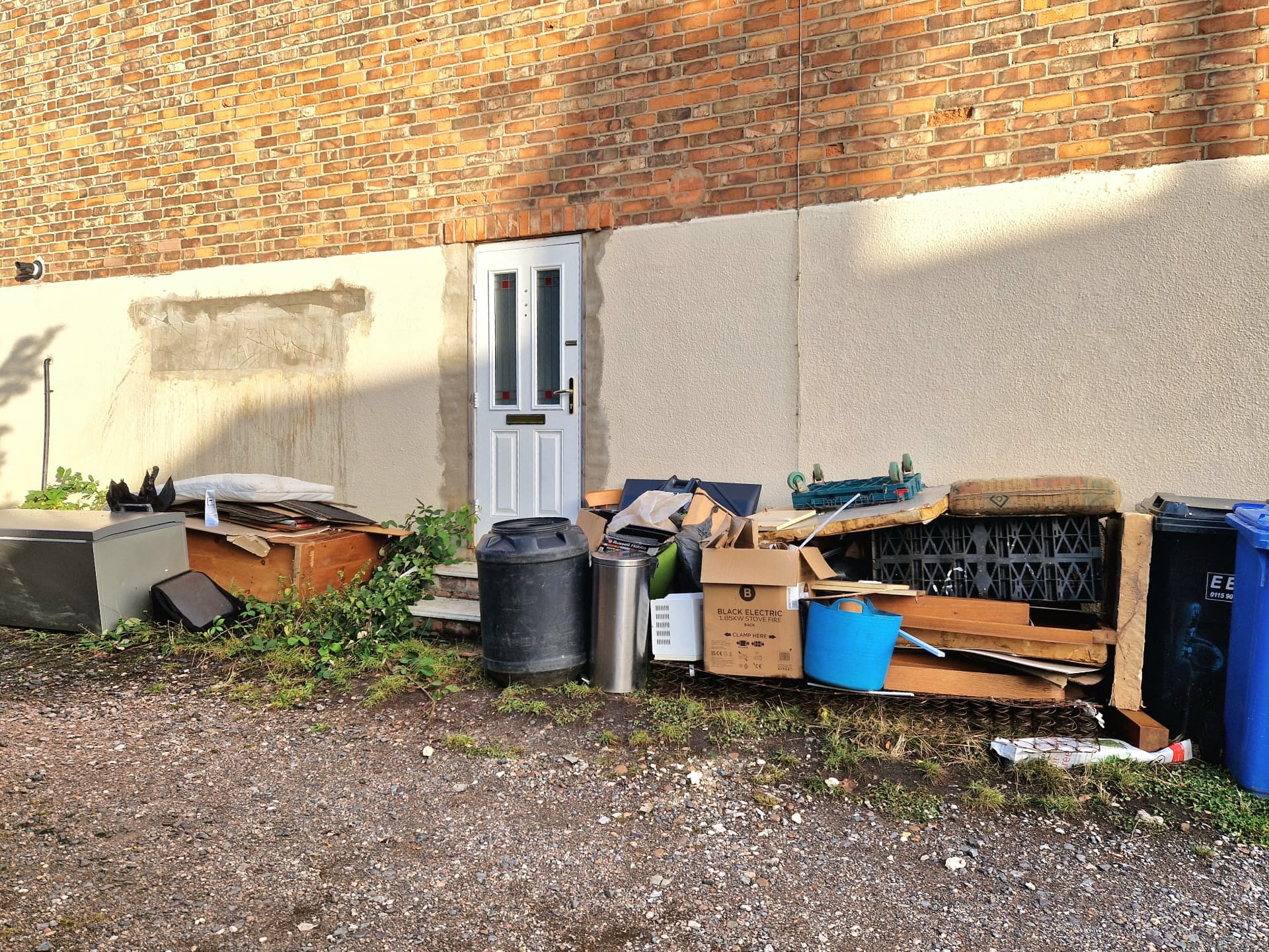 Fly-tipped mattresses, furniture, crates and household rubbish dumped against a wall beside wheelie bins