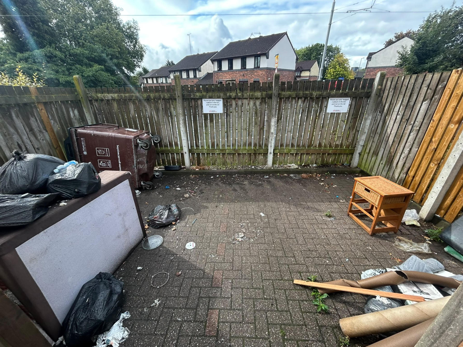 Before house clearance: communal bin store in Manchester filled with black bin bags, a discarded headboard and a bedside table beside a tipped council bin