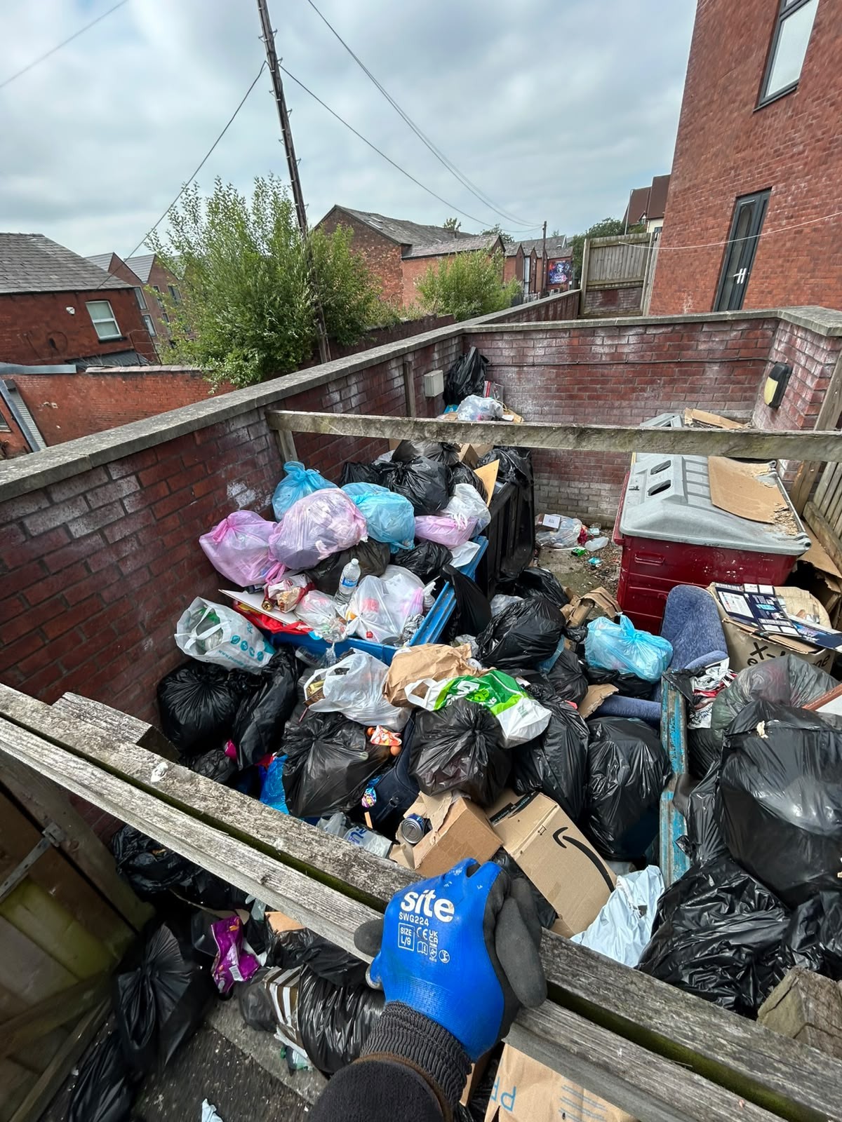 Before waste removal: Bolton communal bin chamber overflowing with bagged household rubbish, cardboard and broken furniture